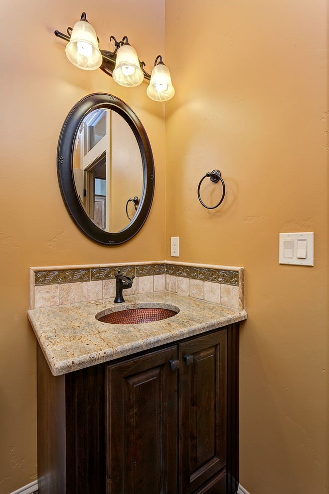 Medium sized mediterranean cloakroom in Boise with raised-panel cabinets, dark wood cabinets, beige tiles, stone tiles, yellow walls, a submerged sink and granite worktops.