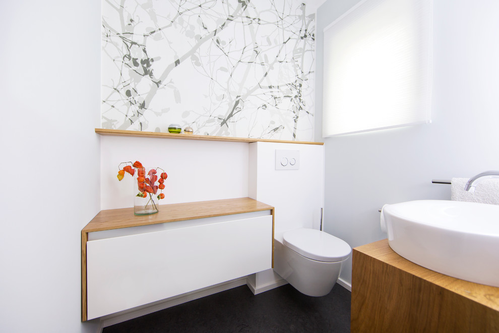 This is an example of a medium sized modern cloakroom in Frankfurt with flat-panel cabinets, white cabinets, a wall mounted toilet, white walls, a vessel sink, wooden worktops and brown worktops.
