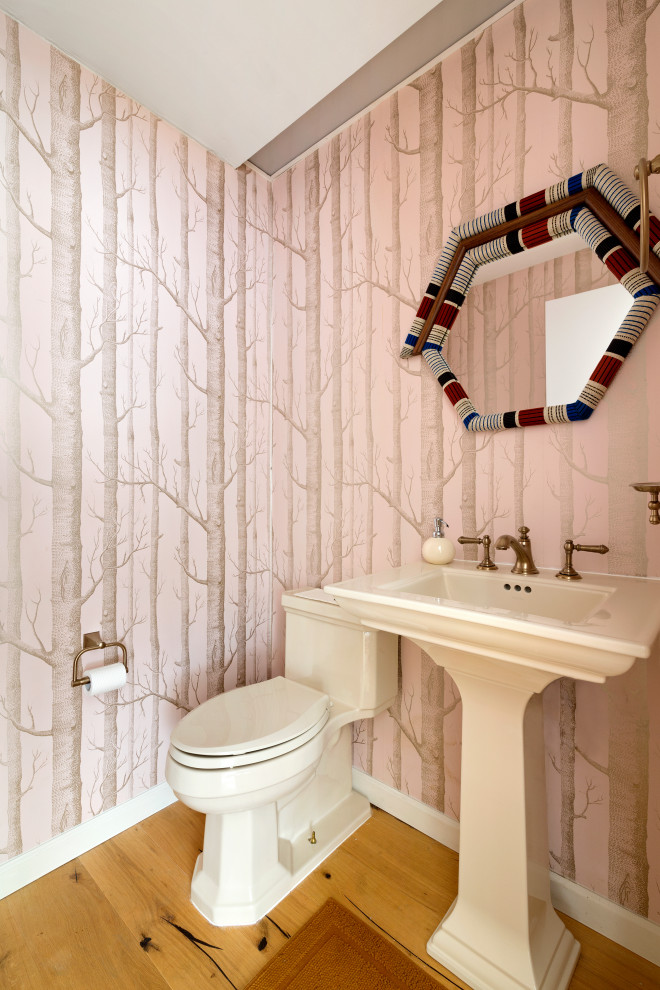 Imagen de aseo contemporáneo con sanitario de una pieza, paredes rosas, suelo de madera en tonos medios, lavabo con pedestal, suelo marrón y papel pintado