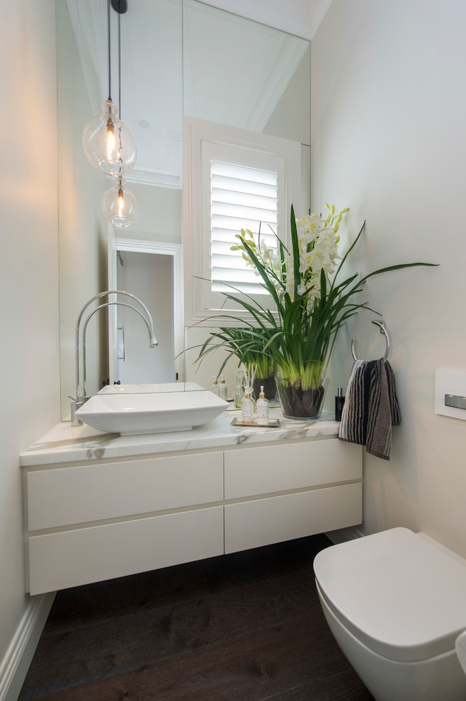 Klassisk inredning av ett litet toalett, med ett fristående handfat, vita skåp, marmorbänkskiva, en vägghängd toalettstol, vita väggar och mellanmörkt trägolv