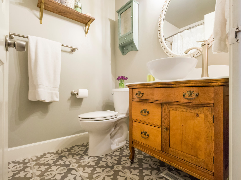 This is an example of a country cloakroom in Boise with freestanding cabinets, medium wood cabinets, a two-piece toilet, beige walls, ceramic flooring, a vessel sink, grey floors and white worktops.