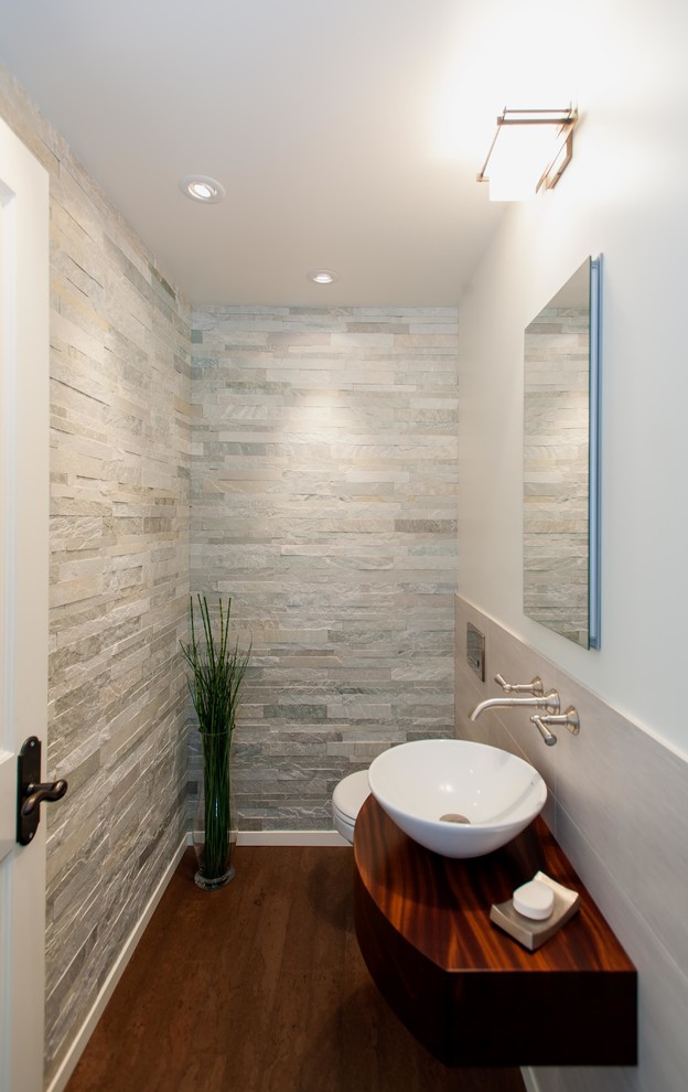 Inredning av ett modernt litet brun brunt toalett, med en vägghängd toalettstol, vita väggar, mellanmörkt trägolv, ett fristående handfat, träbänkskiva och grå kakel