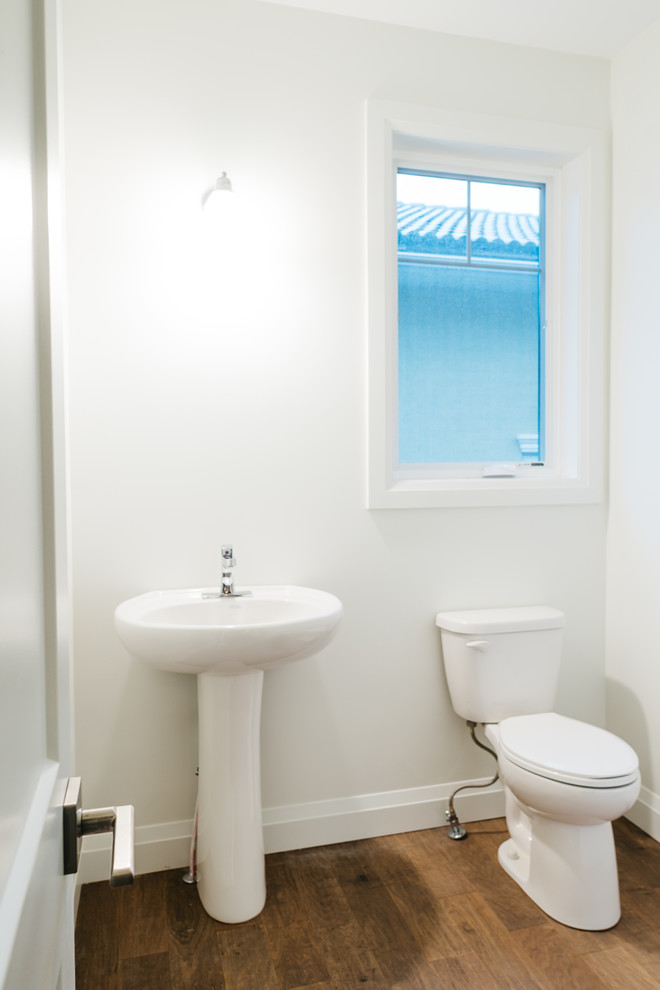 Rustik inredning av ett mellanstort toalett, med en toalettstol med hel cisternkåpa, vita väggar, mellanmörkt trägolv, ett piedestal handfat och brunt golv