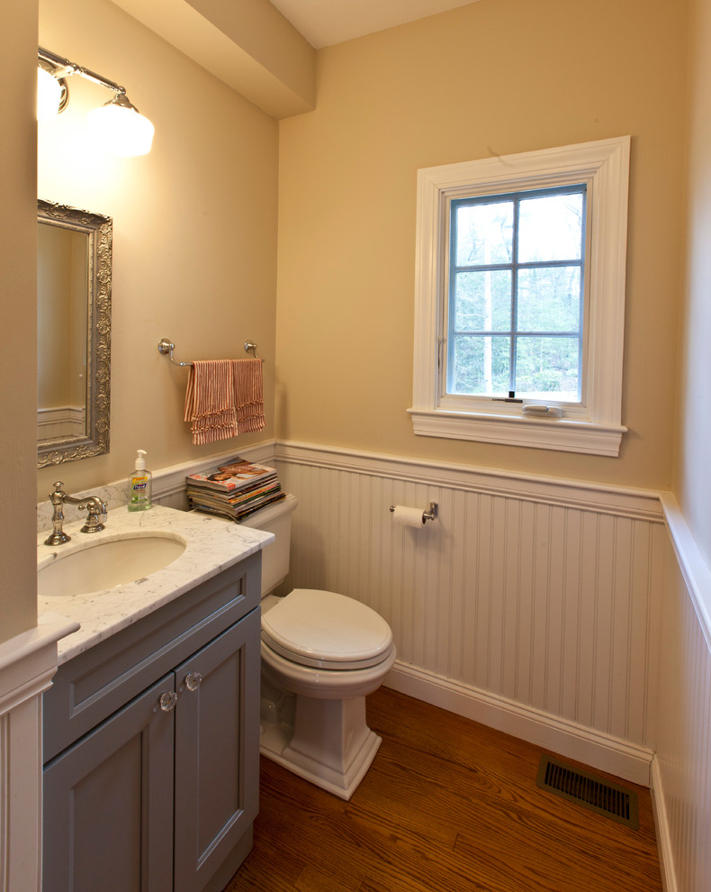 Inspiration for a mid-sized country medium tone wood floor and brown floor powder room remodel in Boston with recessed-panel cabinets, blue cabinets, a two-piece toilet, beige walls, an undermount sink, marble countertops and white countertops