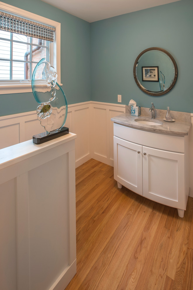 Cette photo montre un WC et toilettes chic avec un placard avec porte à panneau encastré, des portes de placard blanches, un mur bleu, parquet clair, un lavabo encastré et un plan de toilette en marbre.