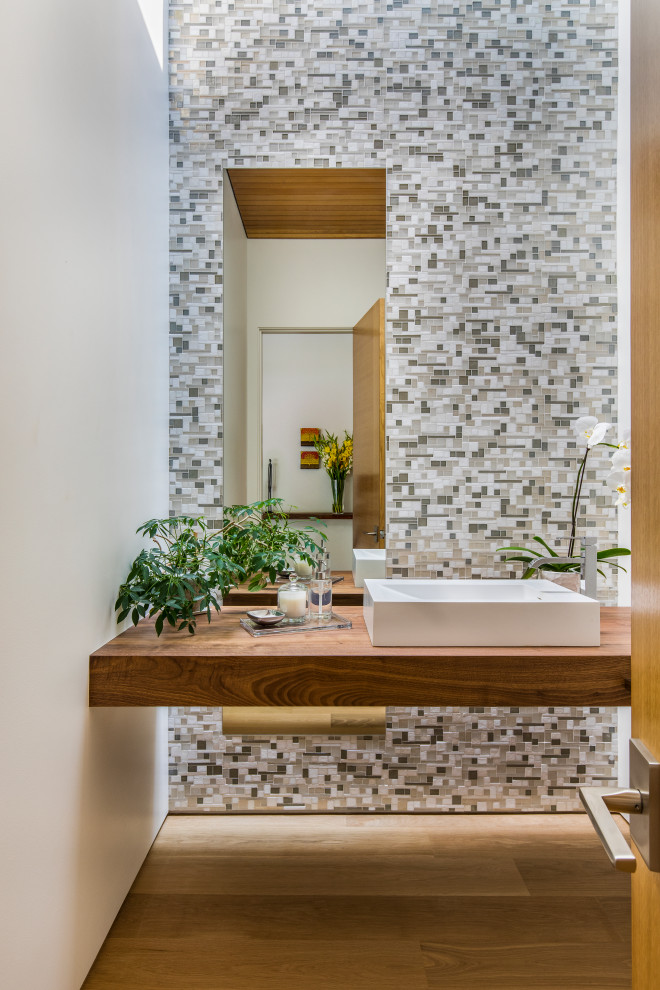 Inredning av ett modernt toalett, med öppna hyllor, skåp i mellenmörkt trä, grå kakel, mosaik, vita väggar, ljust trägolv, ett fristående handfat och träbänkskiva