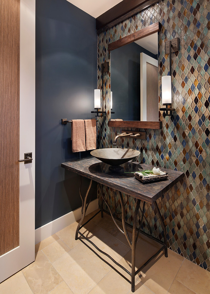 На фото: туалет среднего размера в стиле рустика с бежевой плиткой, синей плиткой, коричневой плиткой, серой плиткой, оранжевой плиткой, разноцветной плиткой, синими стенами, полом из известняка, настольной раковиной, столешницей из дерева и коричневой столешницей