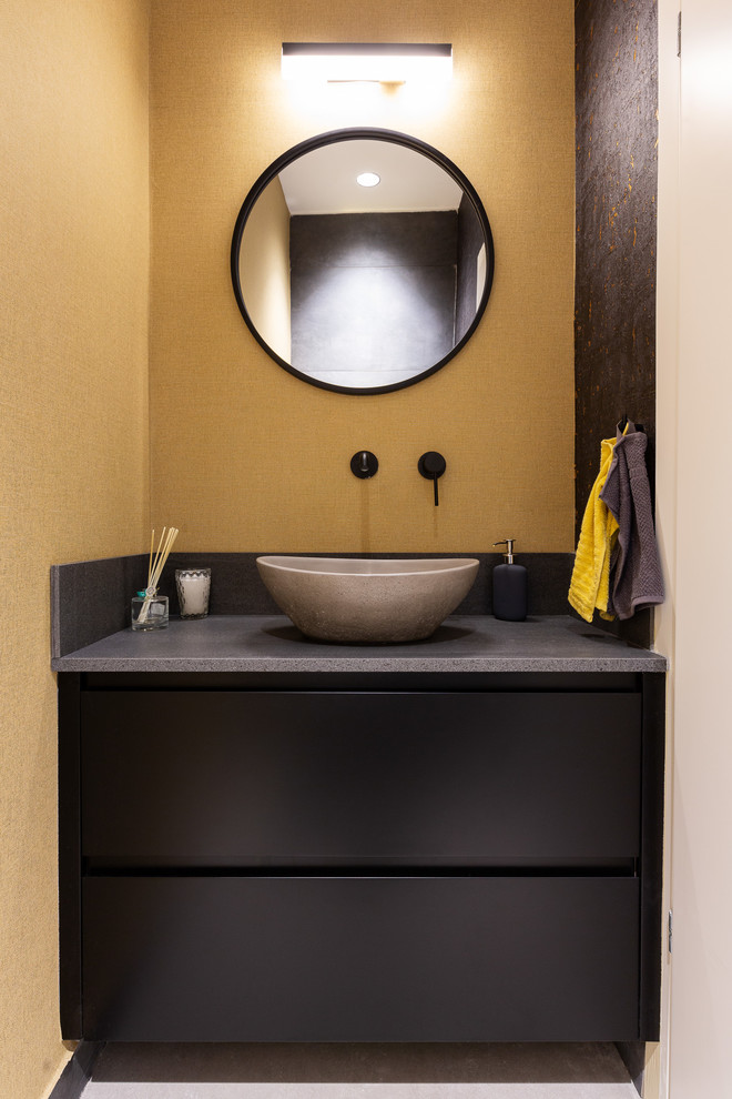 Modelo de aseo retro con armarios con paneles lisos, puertas de armario negras, paredes amarillas, lavabo sobreencimera, suelo gris y encimeras grises