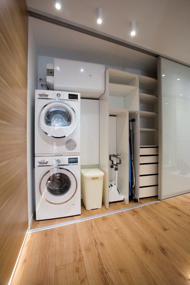 Ejemplo de armario lavadero actual con paredes blancas, lavadora y secadora apiladas y suelo marrón