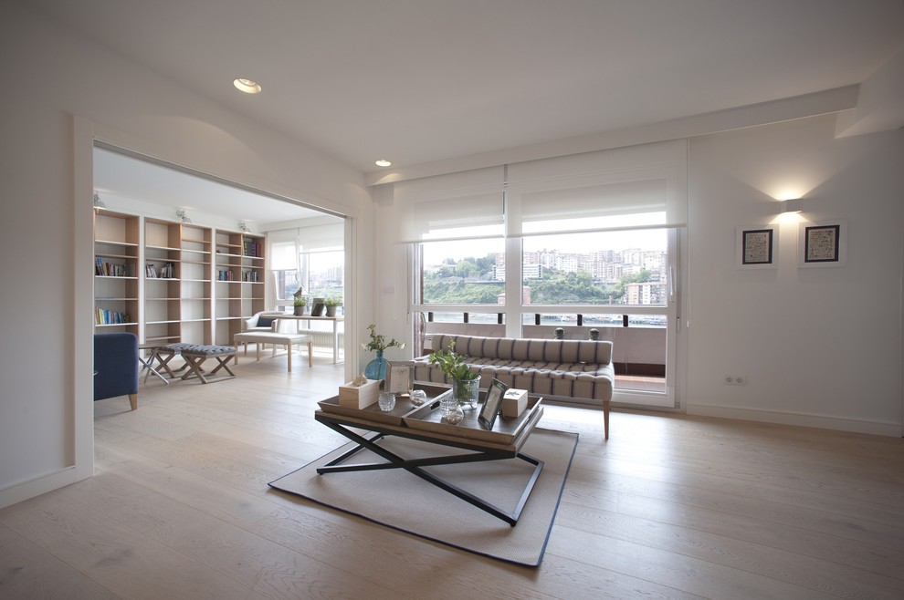 Modelo de sala de estar con biblioteca abierta costera grande sin chimenea y televisor con paredes blancas, suelo de madera clara y suelo marrón