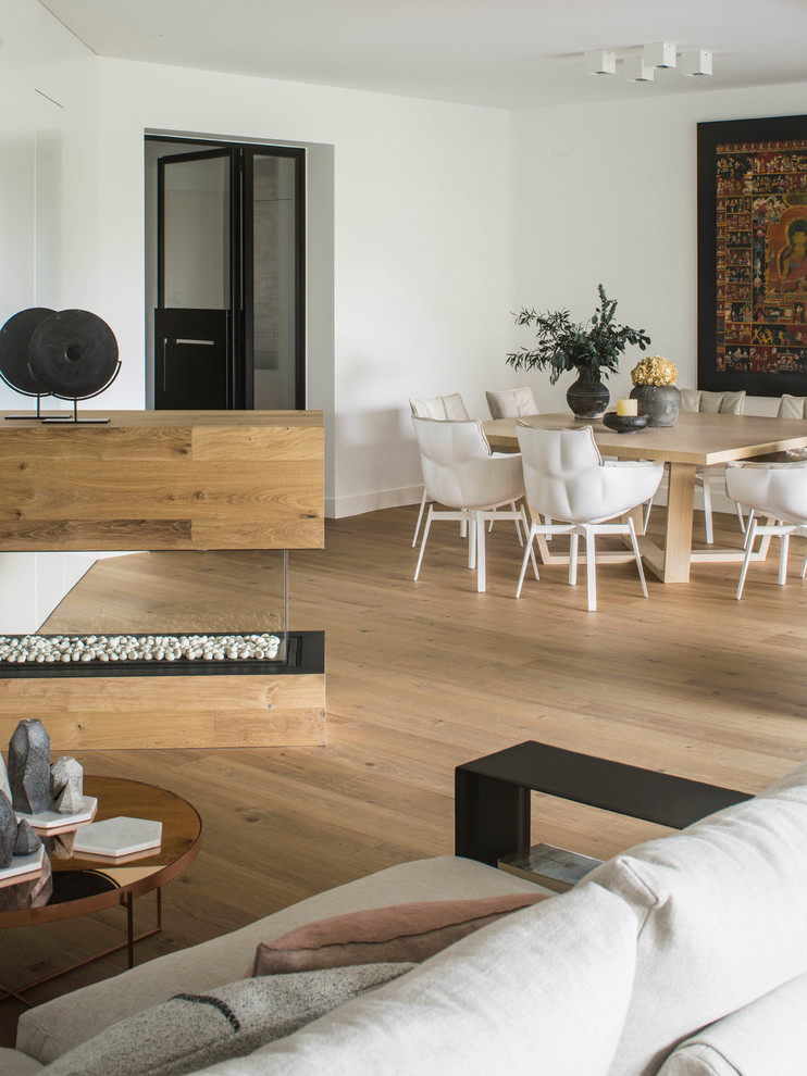 Modelo de sala de estar abierta contemporánea grande con paredes blancas, suelo de madera en tonos medios, chimenea de doble cara y marco de chimenea de madera
