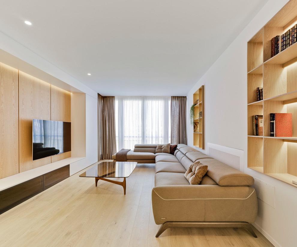 Modelo de sala de estar con biblioteca contemporánea sin chimenea con paredes blancas, suelo de madera clara, televisor colgado en la pared y suelo beige