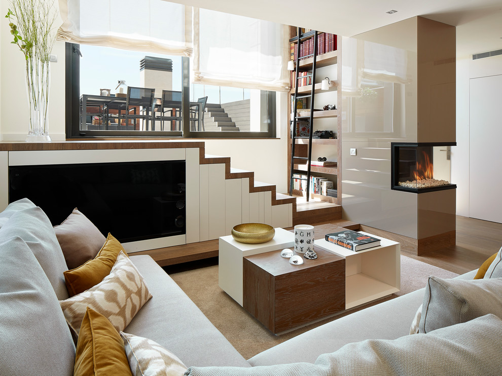 На фото: открытая гостиная комната среднего размера в современном стиле с с книжными шкафами и полками, бежевыми стенами, двусторонним камином, коричневым полом и телевизором на стене с