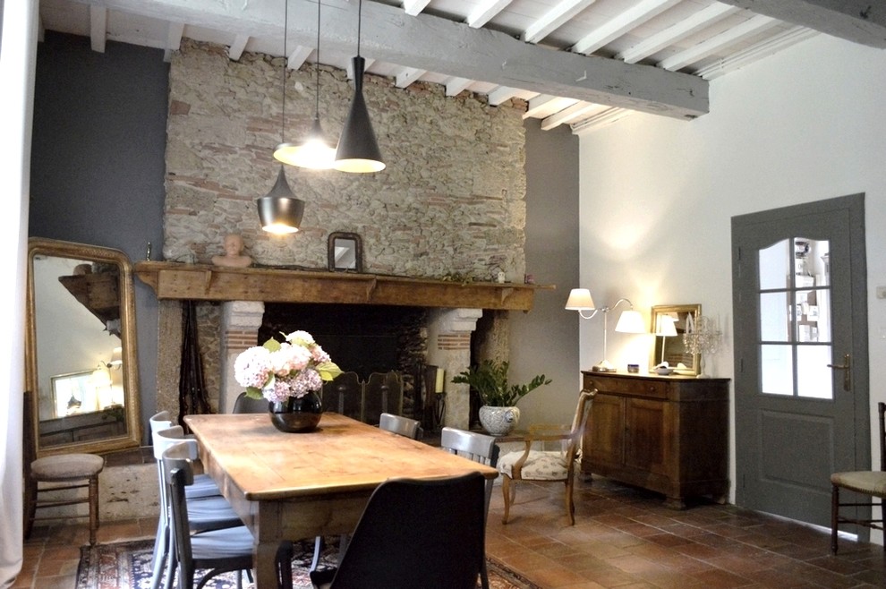 Cette image montre une salle à manger ouverte sur le salon rustique de taille moyenne avec un mur gris, tomettes au sol, une cheminée standard, un manteau de cheminée en pierre et un sol rouge.