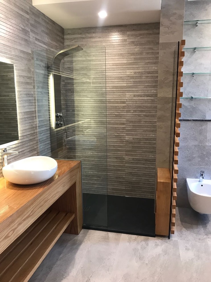Modern inredning av ett mellanstort en-suite badrum, med öppna hyllor, skåp i ljust trä, en kantlös dusch, en vägghängd toalettstol, grå kakel, skifferkakel, grå väggar, skiffergolv, ett nedsänkt handfat, träbänkskiva, grått golv och med dusch som är öppen