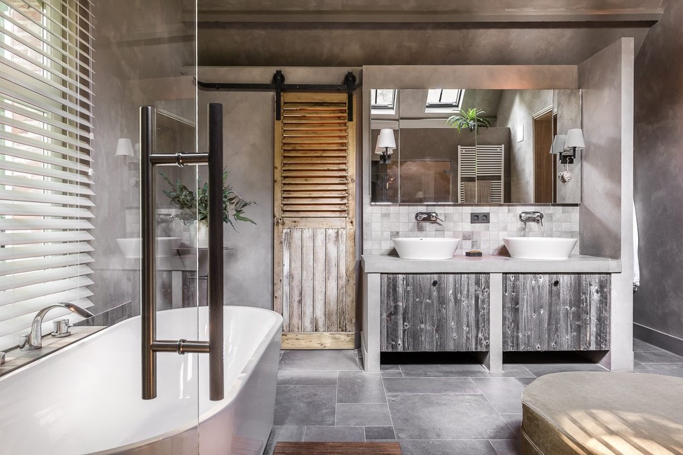 Inredning av ett lantligt en-suite badrum, med släta luckor, skåp i slitet trä, ett fristående badkar, grå väggar, ett fristående handfat, bänkskiva i betong, grått golv och grå kakel