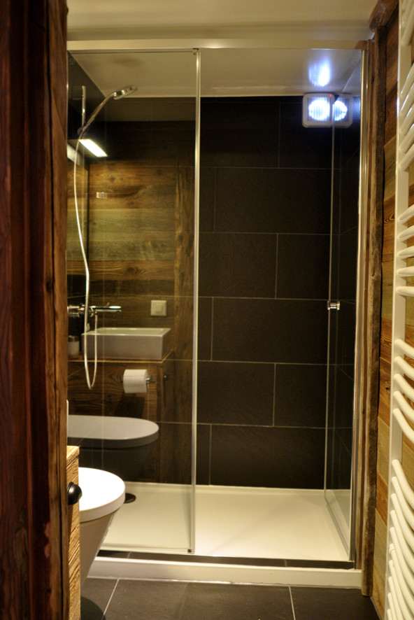 Rustik inredning av ett badrum med dusch, med ett fristående handfat, släta luckor, skåp i slitet trä, träbänkskiva, ett undermonterat badkar, en dubbeldusch, en vägghängd toalettstol, svart kakel, stenhäll, svarta väggar och skiffergolv
