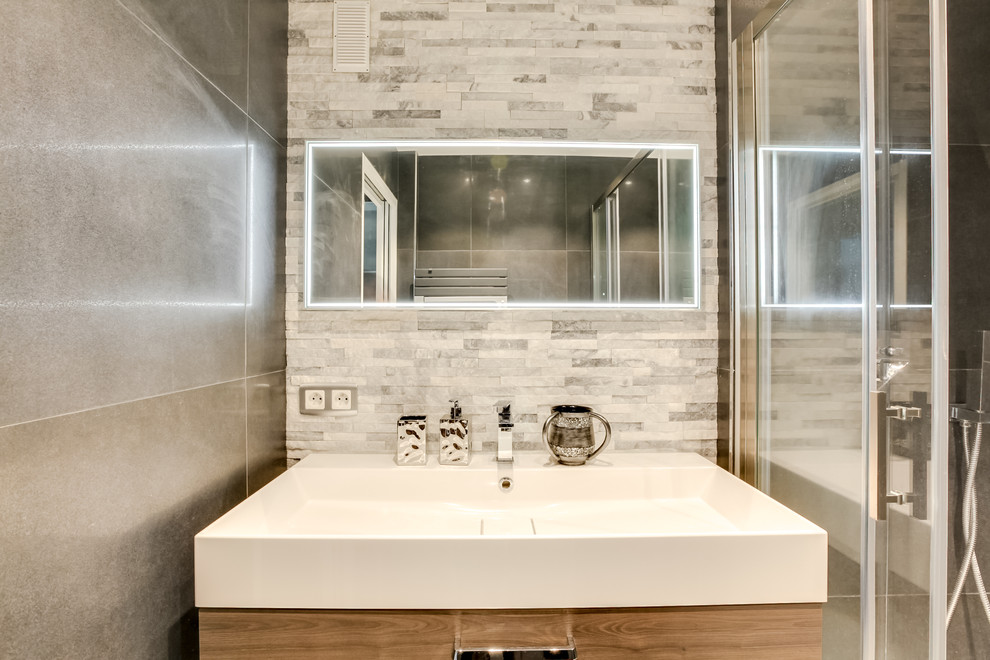 Foto på ett litet funkis badrum med dusch, med skåp i ljust trä, en kantlös dusch, grå kakel, stenhäll, grå väggar, klinkergolv i keramik, bänkskiva i akrylsten, grått golv och dusch med skjutdörr