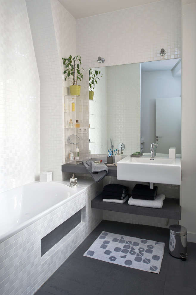 Bild på ett funkis grå grått badrum, med öppna hyllor, bänkskiva i akrylsten, ett platsbyggt badkar, grå kakel, keramikplattor, grå väggar och skiffergolv