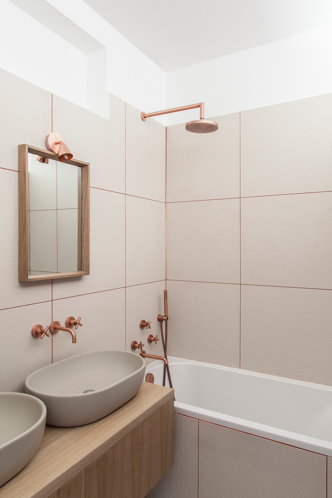 Cette image montre une salle de bain design en bois clair avec un placard à porte plane, une baignoire en alcôve, un combiné douche/baignoire, un carrelage beige, un mur blanc, une vasque et un plan de toilette en bois.