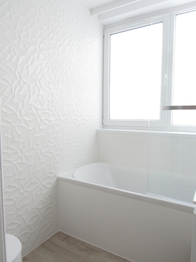 Small contemporary ensuite bathroom in Lille with a submerged bath, a shower/bath combination, a wall mounted toilet, white tiles, ceramic tiles, white walls, light hardwood flooring, an integrated sink and solid surface worktops.