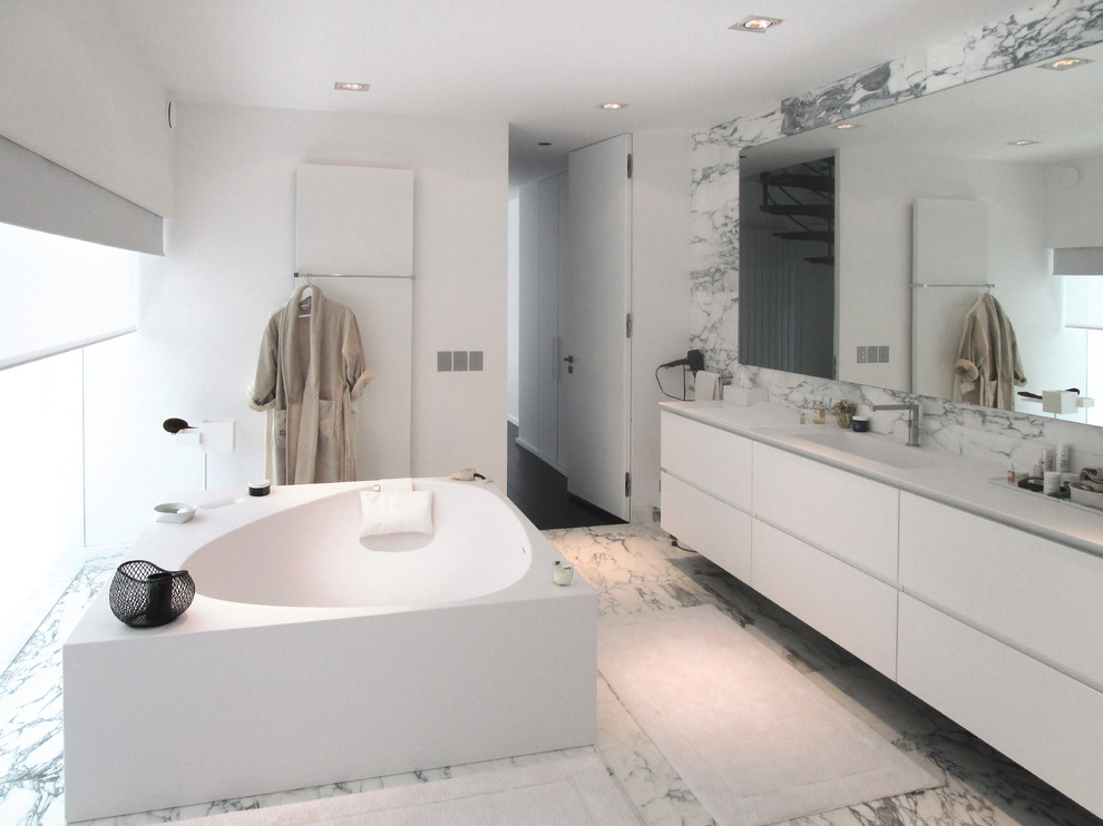 Cette photo montre une grande salle de bain principale tendance avec des portes de placard blanches, une baignoire indépendante, un carrelage de pierre, un mur blanc, un lavabo intégré et un sol en marbre.