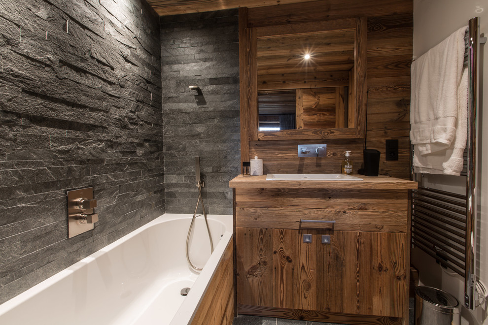 Cette photo montre une salle de bain principale montagne en bois brun avec une baignoire posée, un combiné douche/baignoire, un carrelage de pierre, un lavabo posé, un plan de toilette en bois, un carrelage gris et aucune cabine.