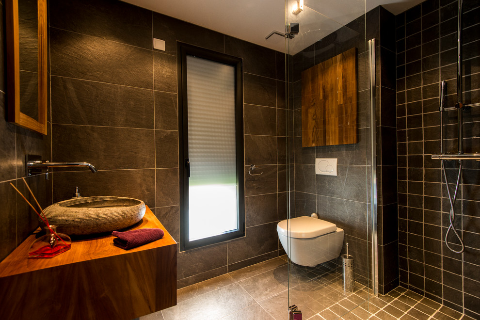 Cette photo montre une salle d'eau tendance de taille moyenne avec WC suspendus, un carrelage noir, un mur noir, un sol en carrelage de céramique, une vasque, un plan de toilette en bois et une douche à l'italienne.