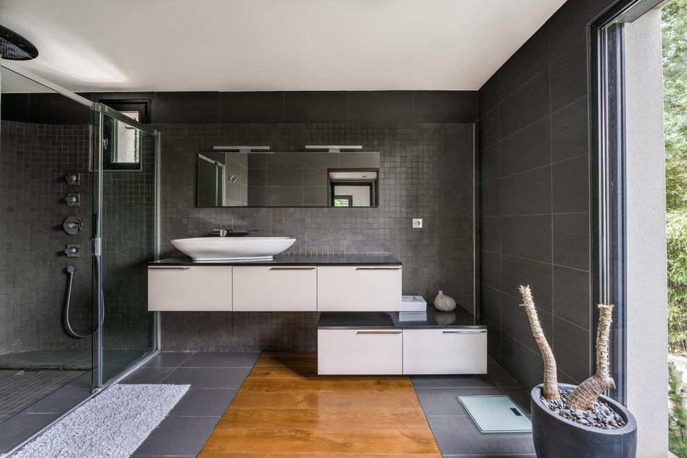 Inspiration pour une salle de bain design avec un placard à porte plane, des portes de placard blanches, un carrelage noir, un carrelage gris, un sol en bois brun, une vasque, un sol marron, une cabine de douche à porte coulissante, un plan de toilette noir et une fenêtre.