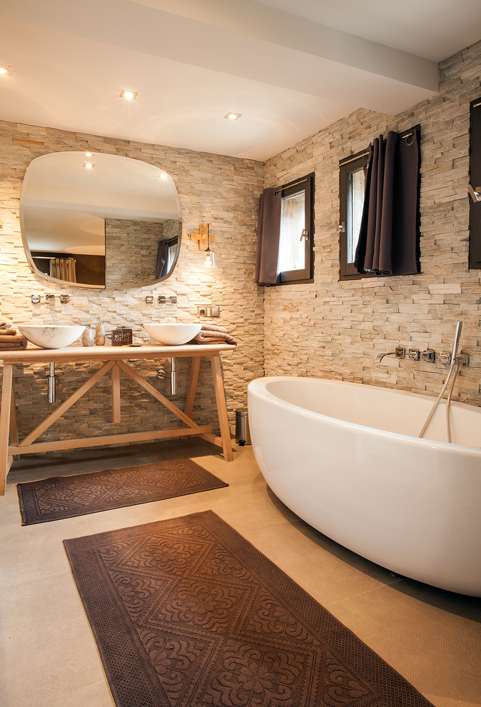 На фото: главная ванная комната в стиле кантри с бежевой плиткой, полом из керамической плитки, столешницей из дерева, отдельно стоящей ванной и настольной раковиной