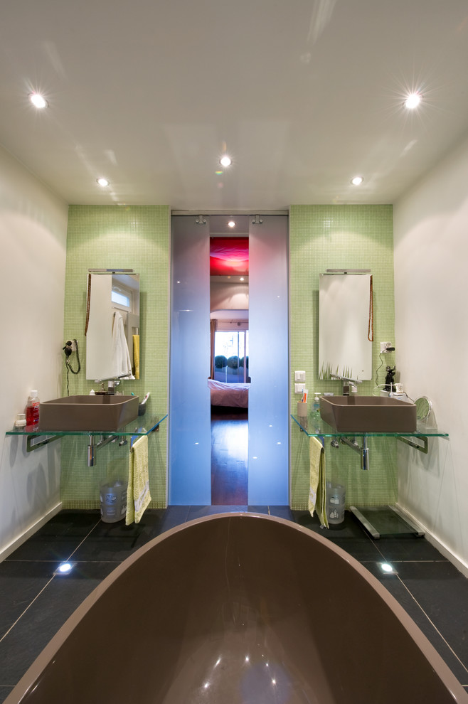 Modelo de cuarto de baño principal actual de tamaño medio con lavabo de seno grande, encimera de vidrio, bañera exenta, ducha a ras de suelo, sanitario de pared, baldosas y/o azulejos verdes, baldosas y/o azulejos en mosaico y paredes blancas