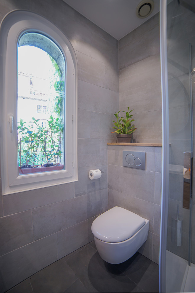 Photo of a small contemporary shower room bathroom in Paris with beaded cabinets, light wood cabinets, a wall mounted toilet, grey tiles, porcelain tiles, grey walls, porcelain flooring, a submerged sink, limestone worktops, grey floors, a hinged door and a corner shower.