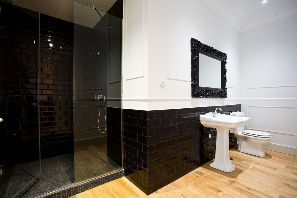 Inredning av ett modernt stort badrum med dusch, med ett piedestal handfat, en dusch i en alkov, svart kakel, vita väggar, ljust trägolv, en toalettstol med hel cisternkåpa och tunnelbanekakel