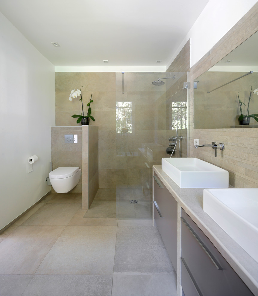 Inredning av ett modernt stort badrum med dusch, med ett fristående handfat, släta luckor, grå skåp, en vägghängd toalettstol, beige kakel, vita väggar, en kantlös dusch och beiget golv