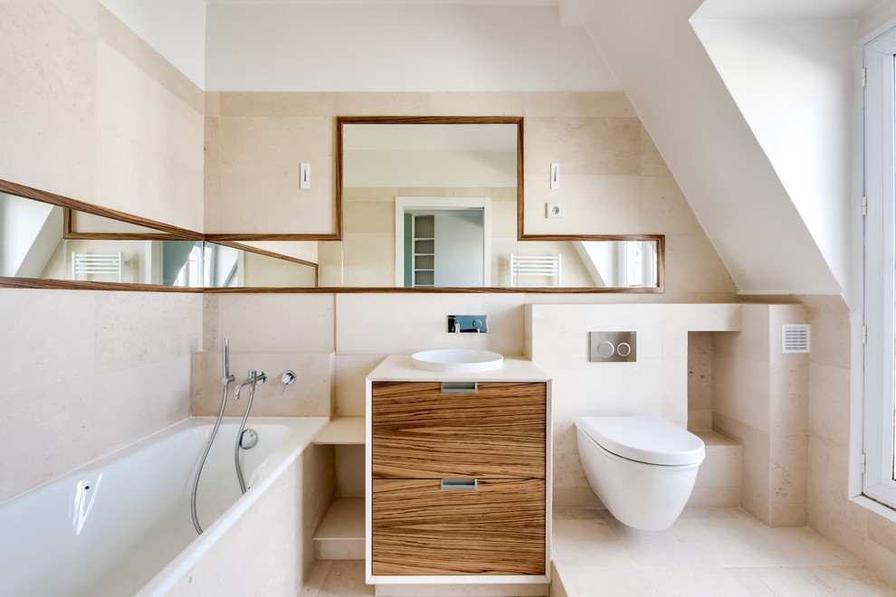 Photo of a large contemporary ensuite bathroom in Paris with a shower/bath combination, a wall mounted toilet, beige tiles, beige walls, marble flooring, a console sink, beige floors, white worktops, medium wood cabinets, a submerged bath, travertine tiles and limestone worktops.