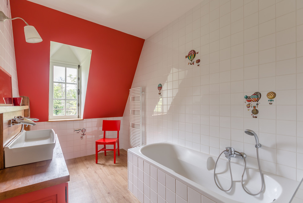 Cette image montre une salle de bain design pour enfant avec une baignoire posée, un combiné douche/baignoire, un carrelage blanc, un mur rouge, parquet clair, une grande vasque, un plan de toilette en bois et un sol beige.