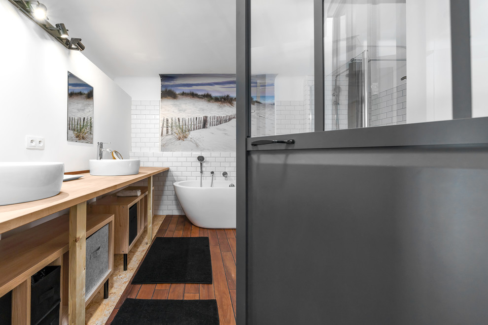 Aménagement d'une salle de bain principale industrielle avec un placard sans porte, une baignoire indépendante, un carrelage blanc, un carrelage métro, un mur blanc, un sol en bois brun et une cabine de douche à porte battante.