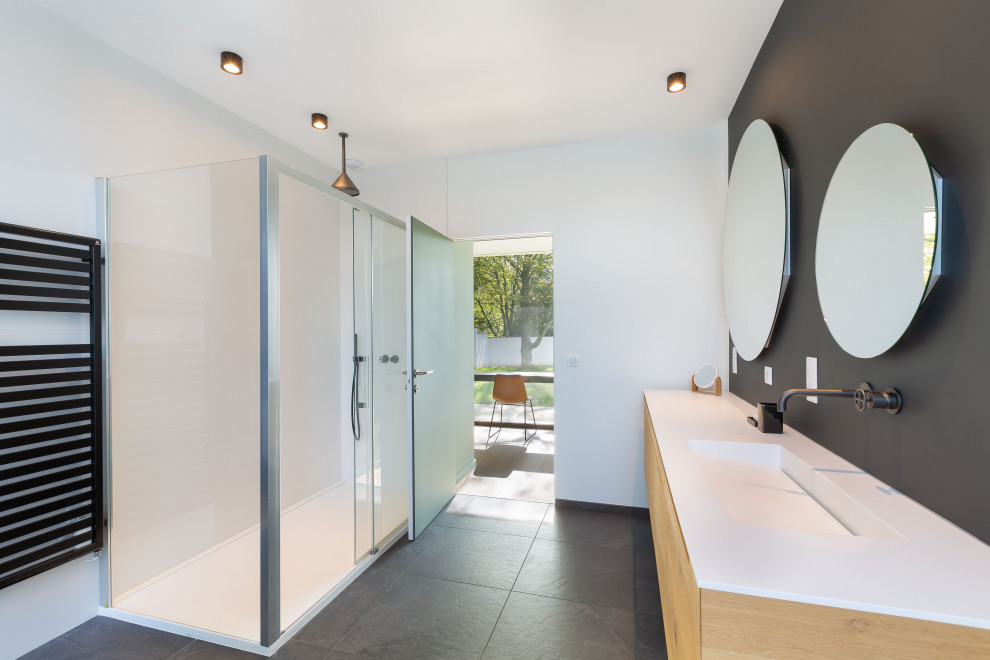 Cette image montre une grande salle de bain principale design en bois clair avec un placard à porte plane, une douche d'angle, un mur blanc, un lavabo intégré, un sol noir, une cabine de douche à porte coulissante, un plan de toilette blanc, meuble double vasque et meuble-lavabo suspendu.