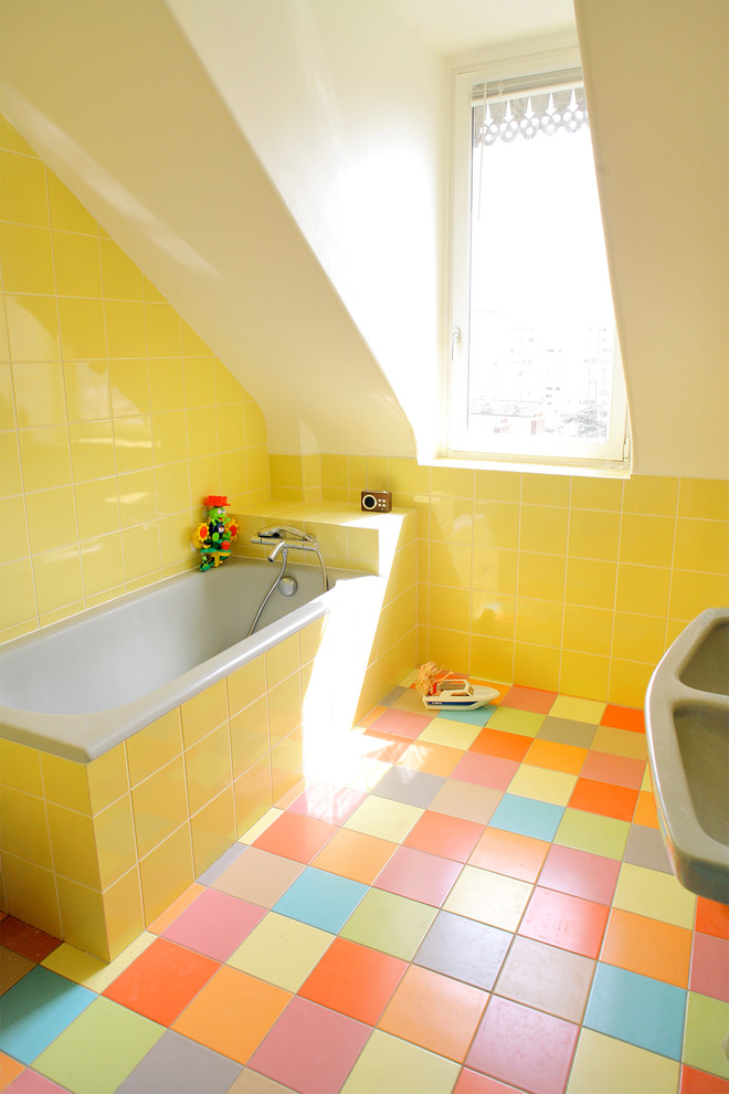 Modern inredning av ett litet badrum för barn, med släta luckor, ett undermonterat badkar, en toalettstol med hel cisternkåpa, gul kakel, porslinskakel, gula väggar, klinkergolv i keramik, ett piedestal handfat och flerfärgat golv