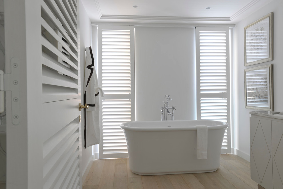 Modelo de cuarto de baño principal clásico renovado de tamaño medio con puertas de armario blancas, bañera exenta, combinación de ducha y bañera, paredes blancas, suelo de madera clara y encimera de mármol