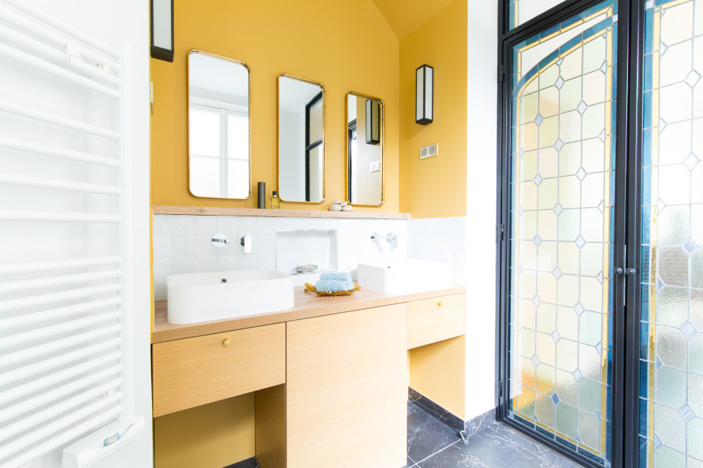 Cette photo montre une grande salle de bain principale tendance en bois clair avec un carrelage blanc, des carreaux de céramique, un mur jaune, un sol en marbre, un plan de toilette en bois, un sol noir, meuble double vasque, un placard à porte plane, une vasque, un plan de toilette beige et meuble-lavabo encastré.