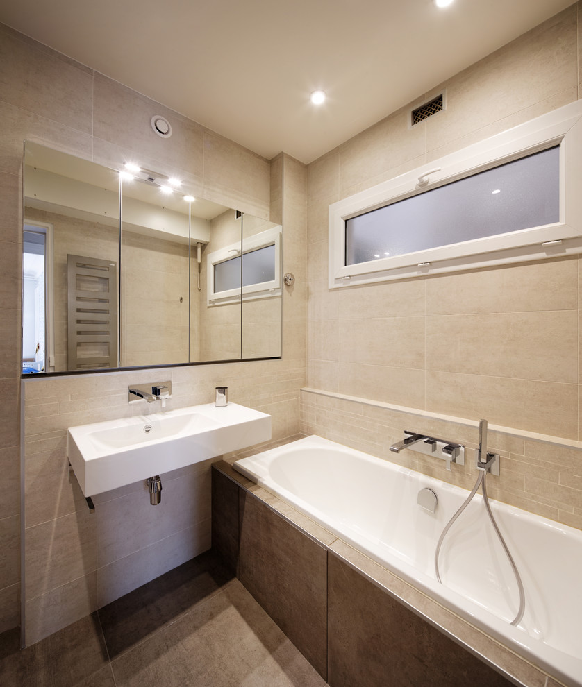 Idée de décoration pour une salle de bain principale design en bois clair de taille moyenne avec un placard à porte affleurante, une baignoire encastrée, un carrelage beige, un carrelage de pierre, un mur beige, un lavabo suspendu et un plan de toilette en surface solide.