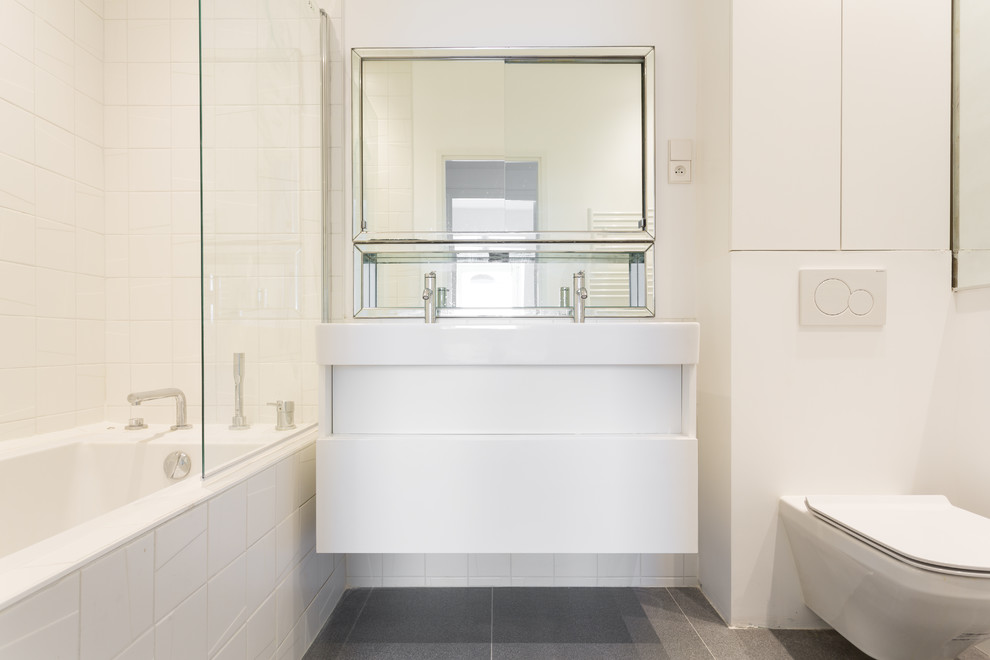 This is an example of a small contemporary ensuite bathroom in Paris with white cabinets, a built-in bath, white tiles, ceramic flooring, grey floors, white worktops, a wall mounted toilet, white walls, flat-panel cabinets, a shower/bath combination, porcelain tiles, a trough sink, solid surface worktops and a hinged door.