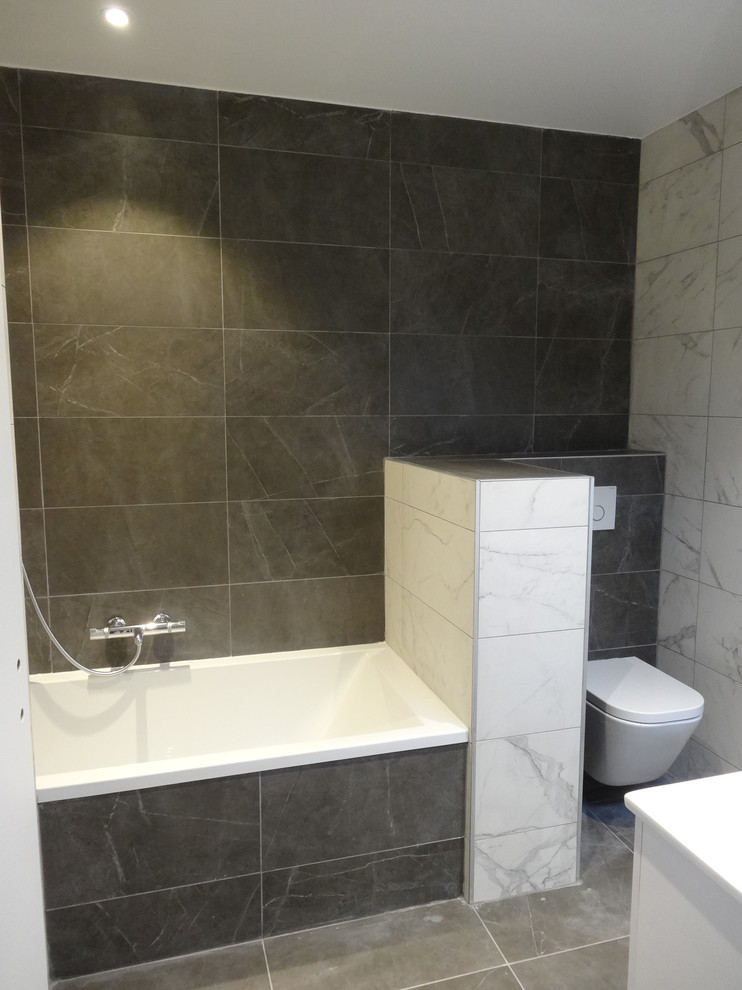 Modern inredning av ett en-suite badrum, med ett hörnbadkar, våtrum, en vägghängd toalettstol, svart kakel, porslinskakel, svarta väggar, klinkergolv i keramik och grått golv