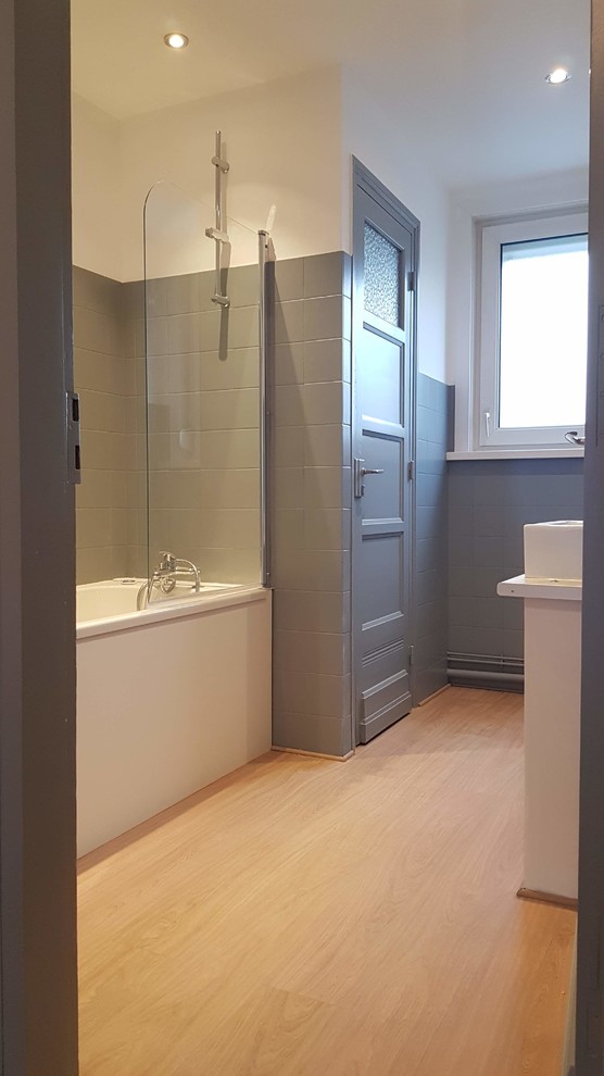 Bild på ett litet funkis badrum, med ett badkar med tassar, våtrum, en toalettstol med separat cisternkåpa, grå kakel, keramikplattor, grå väggar, linoleumgolv och ett nedsänkt handfat