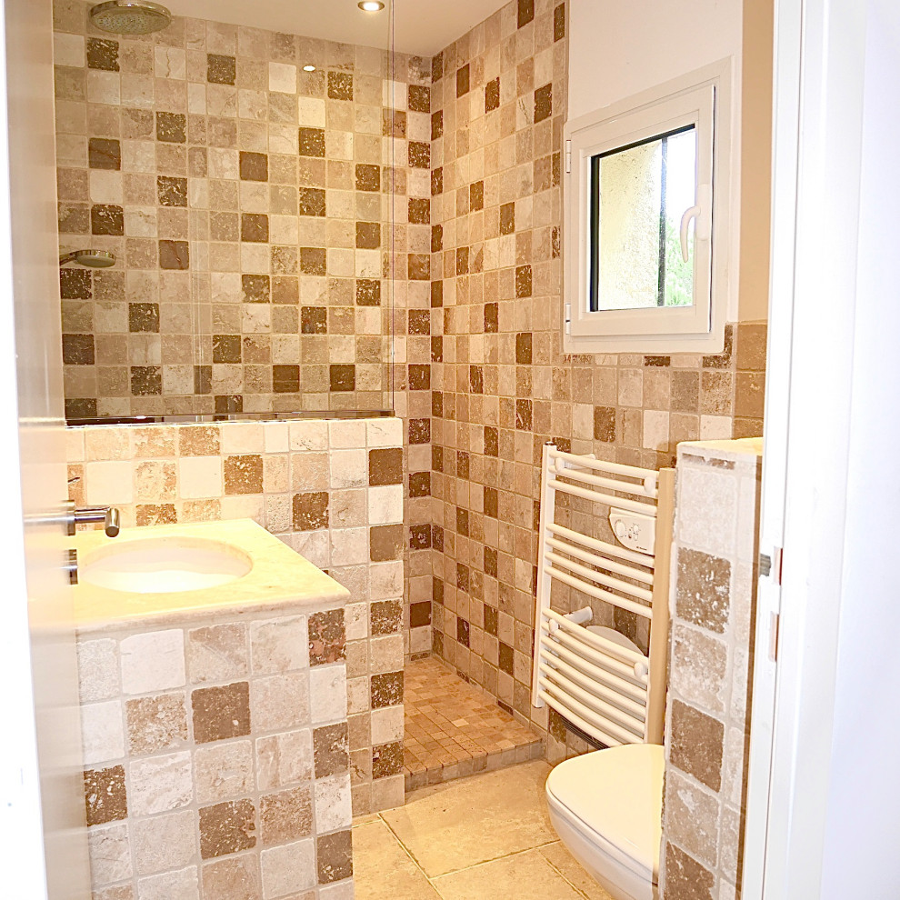 Foto på ett litet lantligt brun badrum med dusch, med en kantlös dusch, en vägghängd toalettstol, beige kakel, travertinkakel, vita väggar, travertin golv, ett konsol handfat, kaklad bänkskiva, beiget golv och med dusch som är öppen