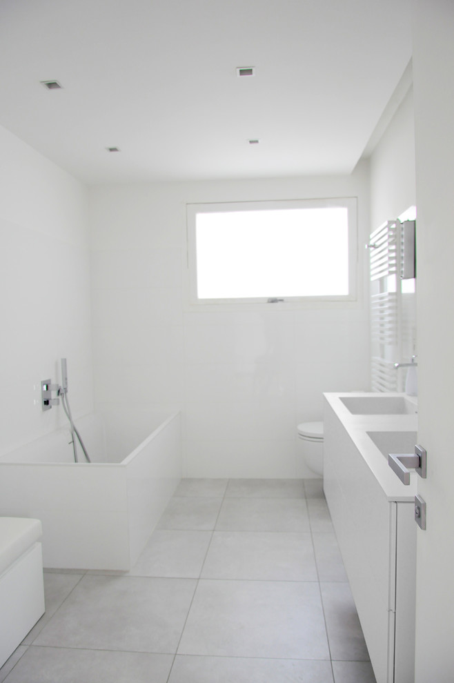Idéer för stora funkis vitt en-suite badrum, med luckor med profilerade fronter, en vägghängd toalettstol, vit kakel, vita väggar, ett konsol handfat, bänkskiva i akrylsten, vita skåp, ett fristående badkar, en öppen dusch, keramikplattor, klinkergolv i keramik, grått golv och med dusch som är öppen