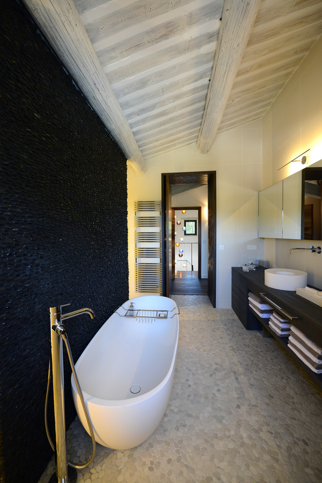 Exemple d'une salle de bain principale tendance de taille moyenne avec une vasque, un placard sans porte, des portes de placard noires, une baignoire indépendante, un sol en galet, un carrelage noir, un mur blanc et des carreaux de céramique.