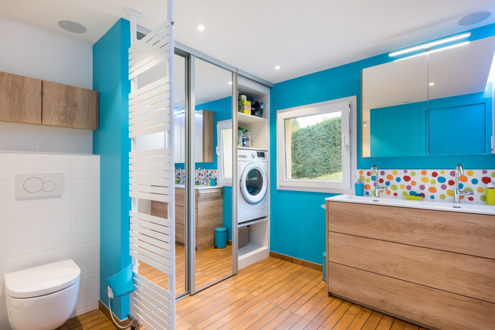 This is an example of a medium sized ensuite bathroom in Lyon with flat-panel cabinets, light wood cabinets, a built-in shower, a wall mounted toilet, multi-coloured tiles, blue walls, wood-effect flooring, a console sink, brown floors, an open shower, white worktops, a laundry area, double sinks, a freestanding vanity unit and a drop ceiling.