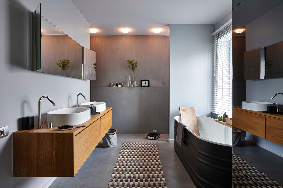 Foto på ett mellanstort funkis en-suite badrum, med släta luckor, skåp i mellenmörkt trä, ett fristående badkar, grå väggar, betonggolv, ett fristående handfat, grå kakel och träbänkskiva